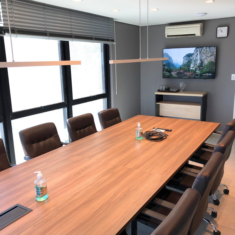 Na foto mostra uma sala de reuniões com uma mesa grande com 10 cadeiras confortáveis e de rodinhas, com hdmi para uso na TV e uma janela em forma de L em toda a extensão da sala.